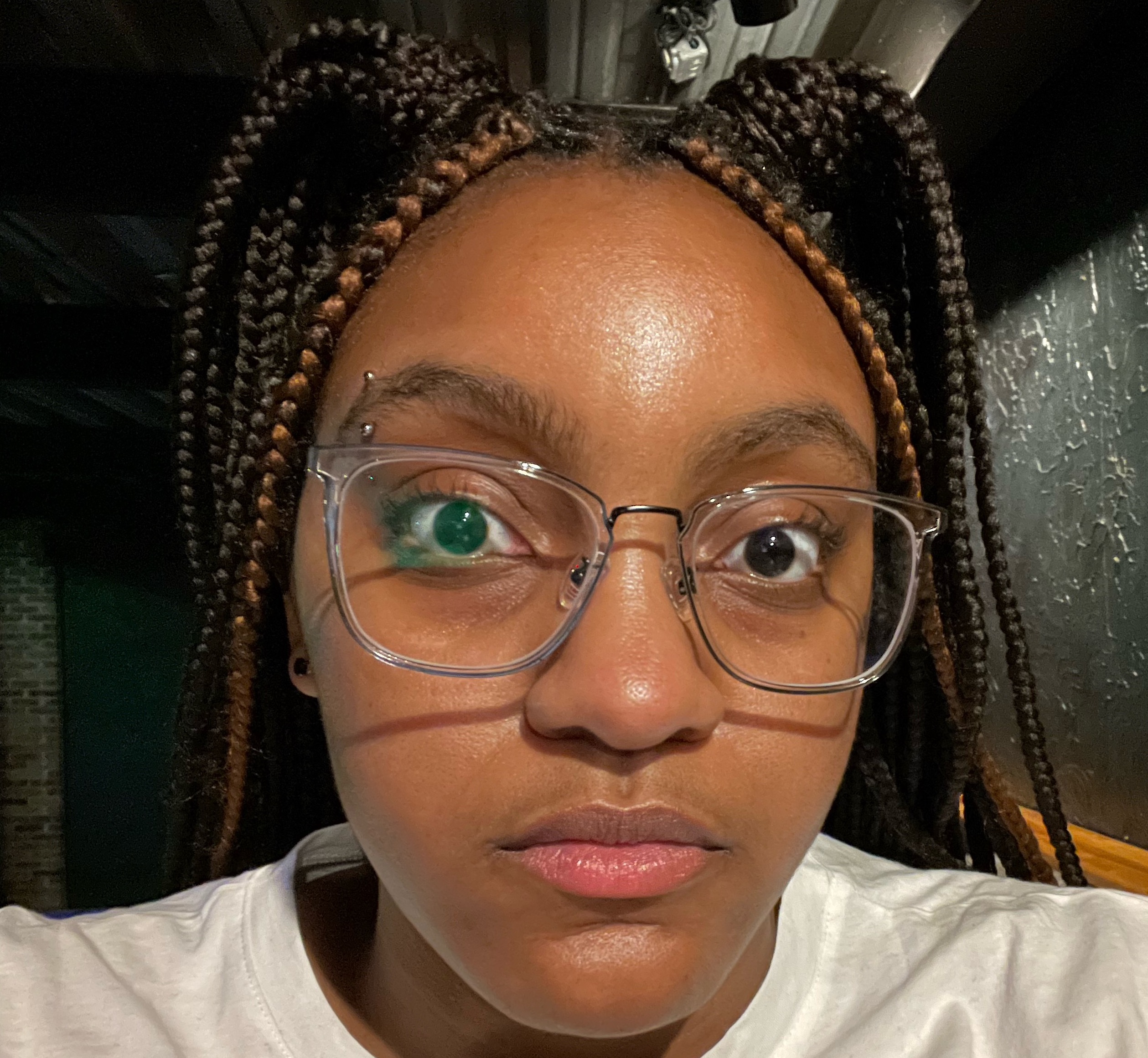 A young African American woman (20 years old) with box dark brown braids with two honey brown highlights in the front. She is wearing clear-rimmed glasses and has a silver barbell eyebrow piercing in her right eyebrow. She has a calm, relaxed facial expression.