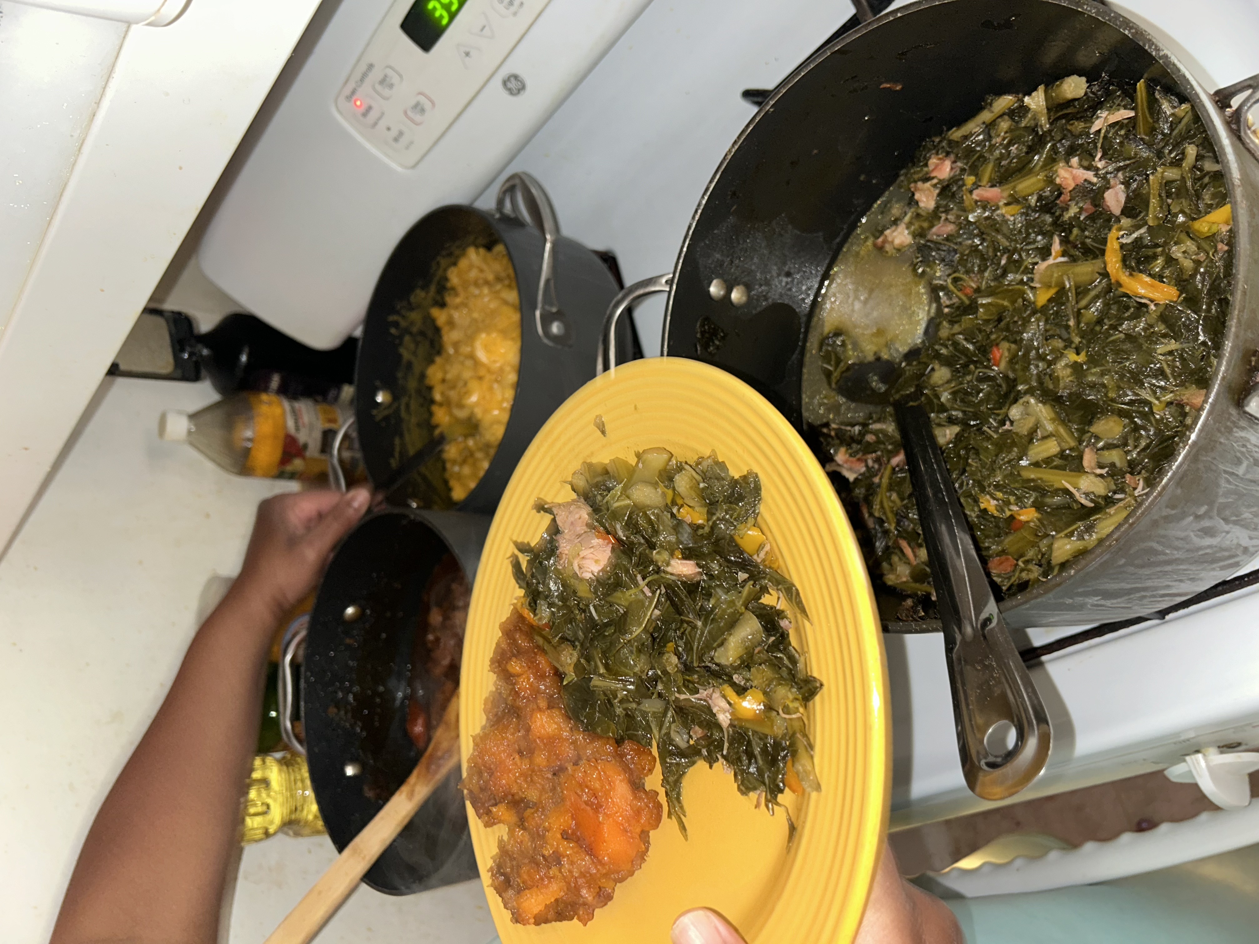 A person holding a yellow plate of food (sweet potatoes and collard greens), reaches into a pot of macaroni and cheese. The macaroni and cheese pot sits on top of a white stove along with a sweet potato and collard green pot as well.