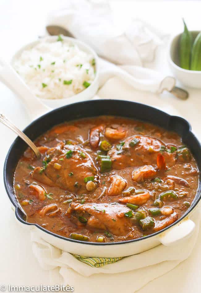 A white pot of gumbo soup with a silver spoon. Behind the bowl of gumbo is a small white bowl filled with white rice and chives.