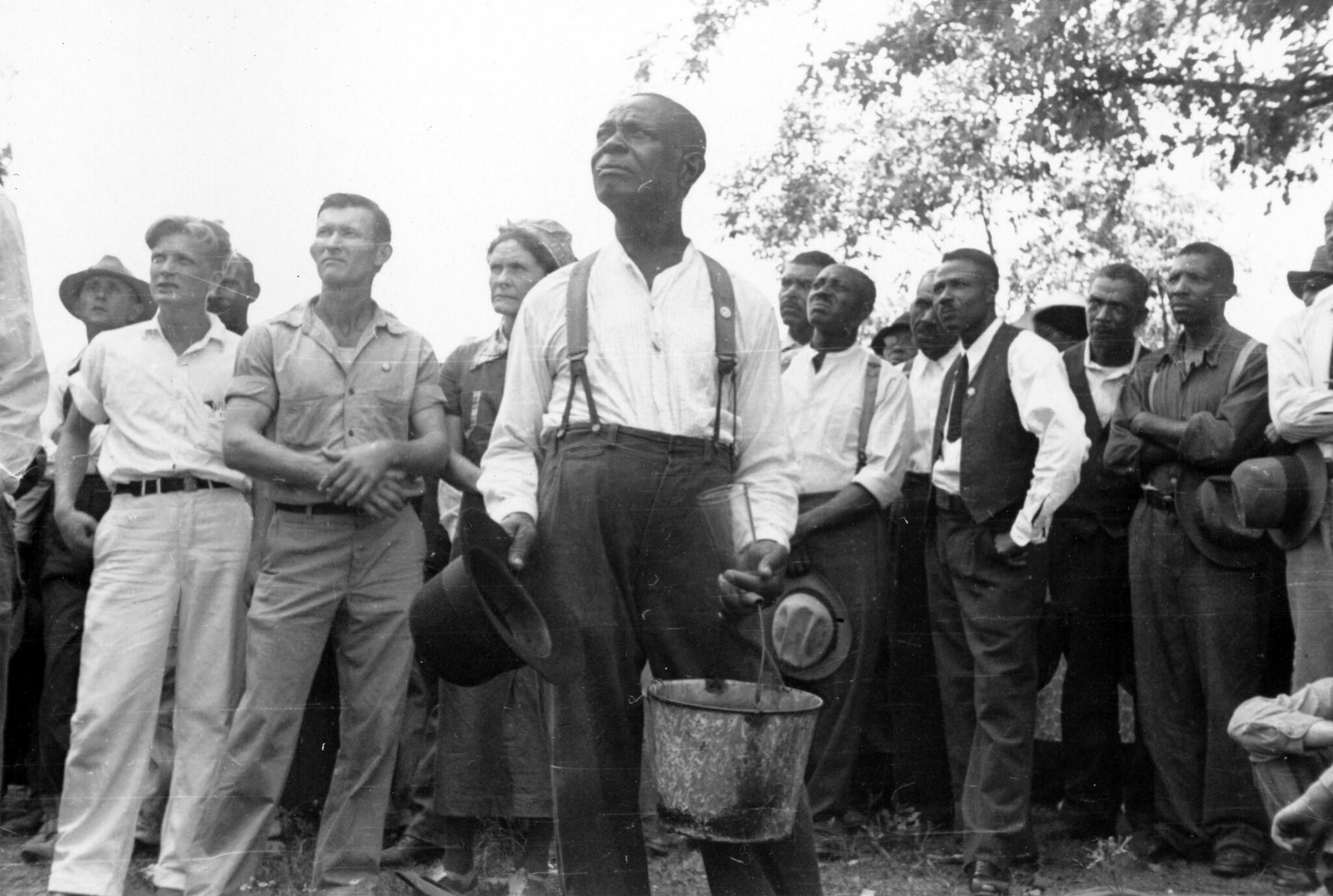 A black and white photograph depicting a group of men standing all facing in one direction on a grassy field outside. They are both white and black men and the setting is 19th century South of the United States. The men are wearing suits, belts, and are carrying hats.